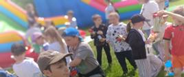 Grupa dzieci stoi na trawie podczas festynu lub pikniku szkolnego. NW tle widoczny kolorowy dmuchany zamek.