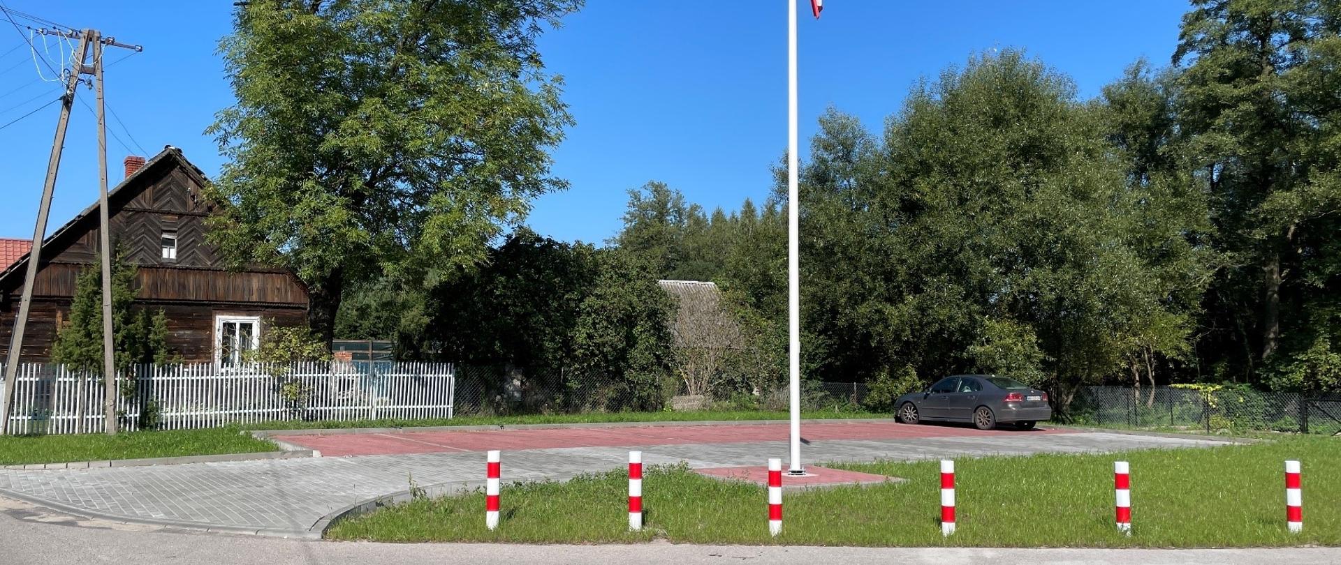 Kilkustanowiskowy parking na którym zaparkowano samochód. Znajduje się tu również maszt z białoczerwoną flagą. Wokół drzewa oraz dom z ogrodzeniem.