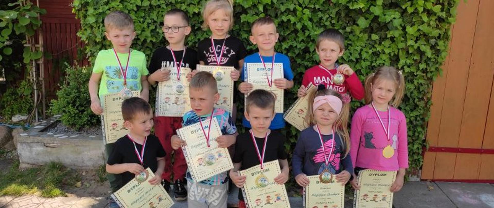 Grupa dziesięciu dzieci stoi na tle ściany pokrytej zieloną rośliną. Na szyjach mają medale, w rękach dyplomy. 
