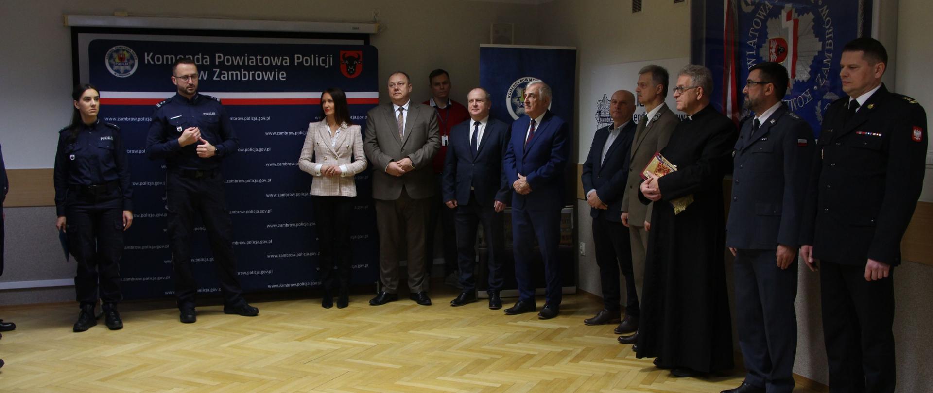 Komendant Daniel Szymanowicz składa świąteczne życzenia do zgromadzonych: Funkcjonariusze policji w ciemnogranatowych mundurach, mężczyźni w garniturach oraz kobieta w jasnym żakiecie, duchowny w czarnej sutannie, trzymający Pismo Święte, mężczyzna w mundurze leśnika.