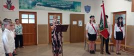Pani Dyrektor stoi na środku i przemawia przez mikrofon. Po prawej stronie znajduje się troje uczniów - dwie dziewczyny i chłopak. Wszyscy ubrani są w stroje galowe. Chłopak trzyma w dłoni sztandar szkoły.