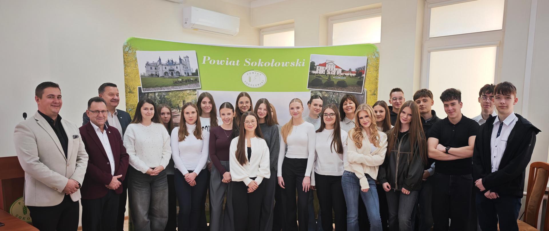Uczniowie wraz ze starostą i pozostałymi uczestnikami pozują do grupowego zdjęcia na tle ścianki w sali konferencyjnej Starostwa Powiatowego w Sokołowie Podlaskim