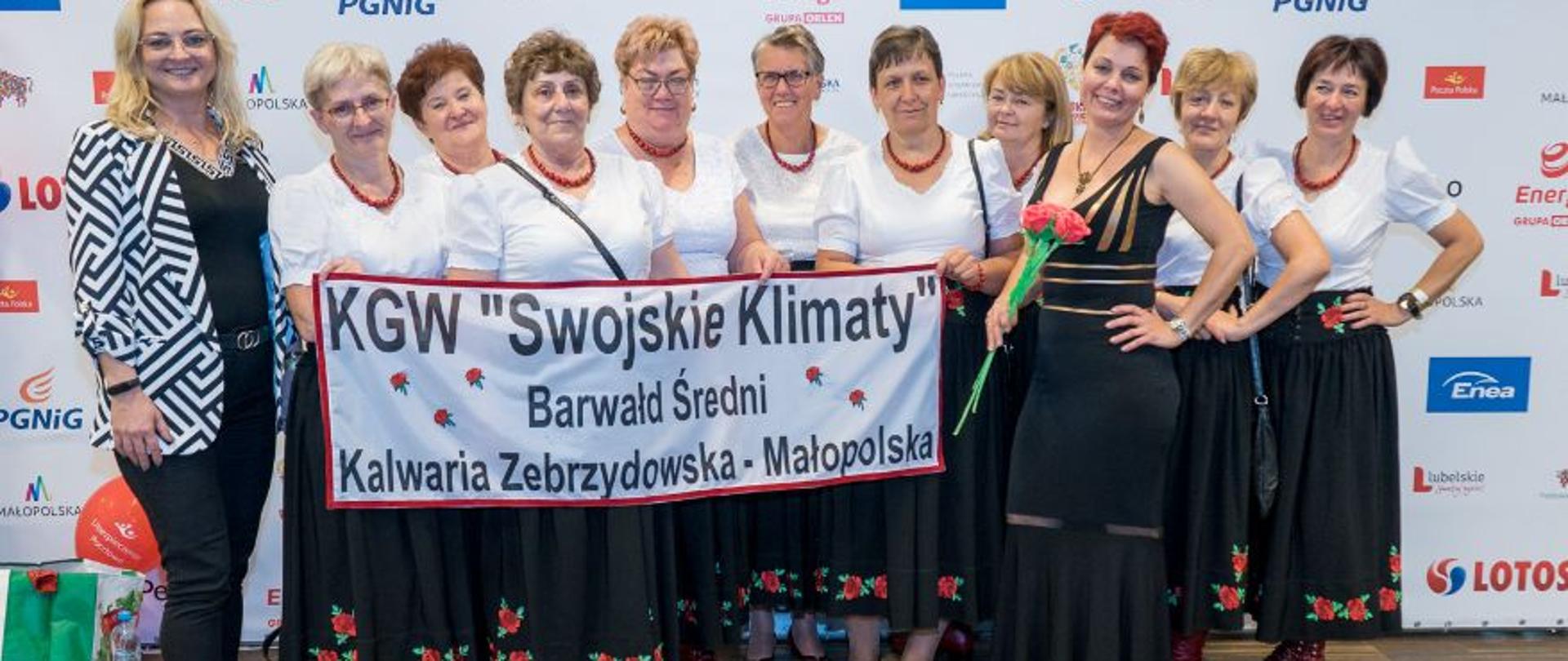Zdjęcie grupowe KGW „Swojskie Klimaty”, na zdjęciu przedstawicielki koła z banerem o treści KGW "Swojskie Klimaty" Barwałd Średni - Kalwaria Zebrzydowska - Małopolska. 