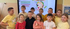 Uczniowie z klasy 3 stoją przed tablicą interaktywną, na której wyświetlona jest informacja o uzyskaniu przez klasę tytułu laureata w konkursie