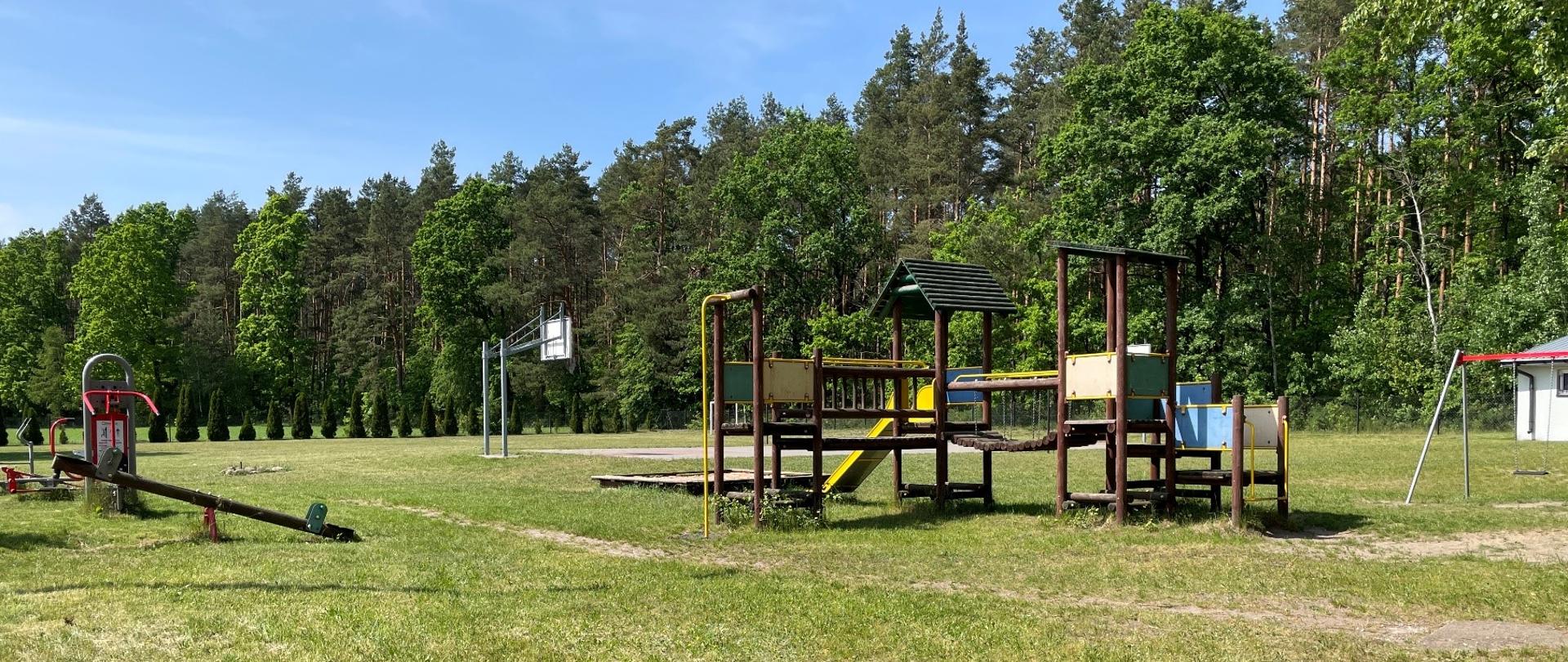 Plac zabaw, siłownia plenerowa oraz boisko do koszykówki. W tle las.