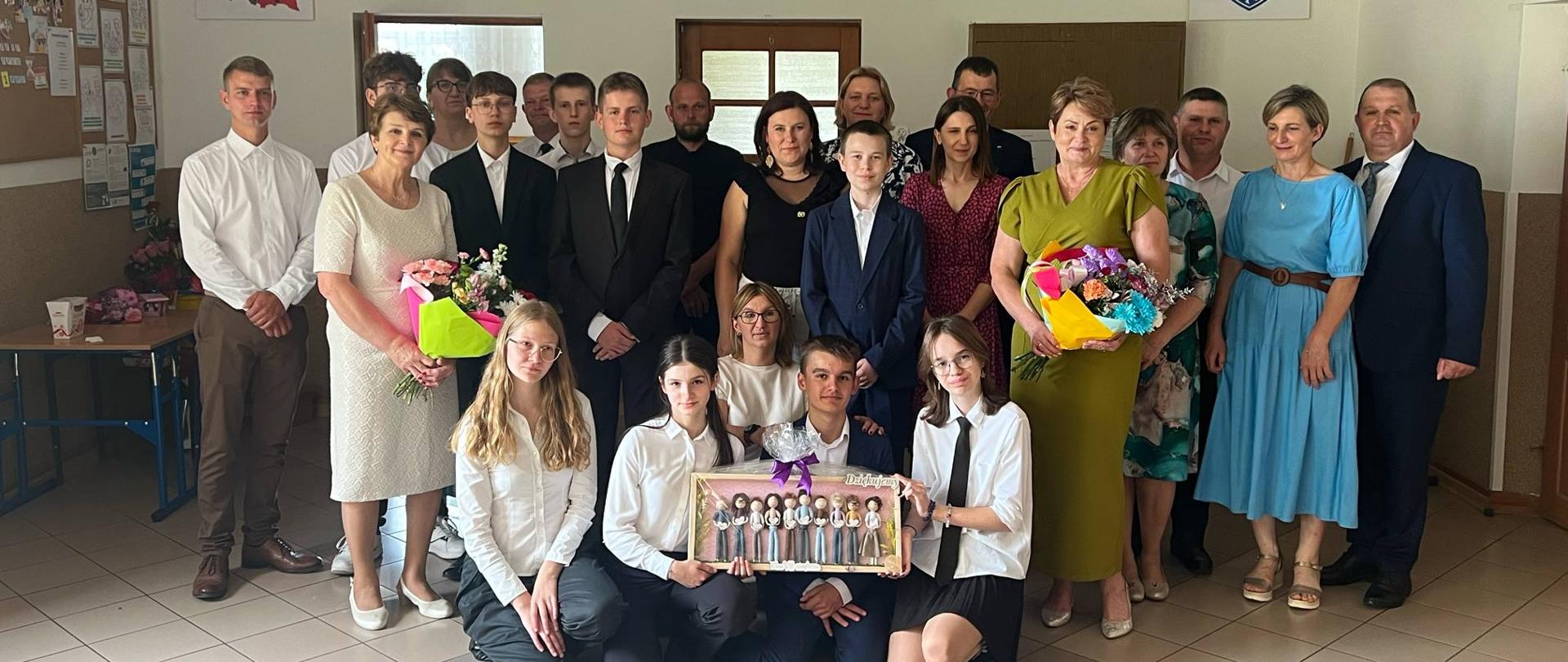 Zdjęcie grupowe uczniów klasy ósmej ubranych w stroje galowe, ich rodziców, wychowawczyni oraz Pani Dyrektor. Pani Dyrektor oraz wychowawczyni trzymają w dłoniach bukiety z kolorowych kwiatów. 