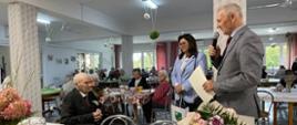 Mężczyzna w szarym garniturze przemawia do mikrofonu, trzymając dokument. Obok stoi kobieta w błękitnym żakiecie, a na wprost nich na wózku inwalidzkim siedzi starszy mężczyzna. Na stole przed nimi ułożone są owoce i ciasta.