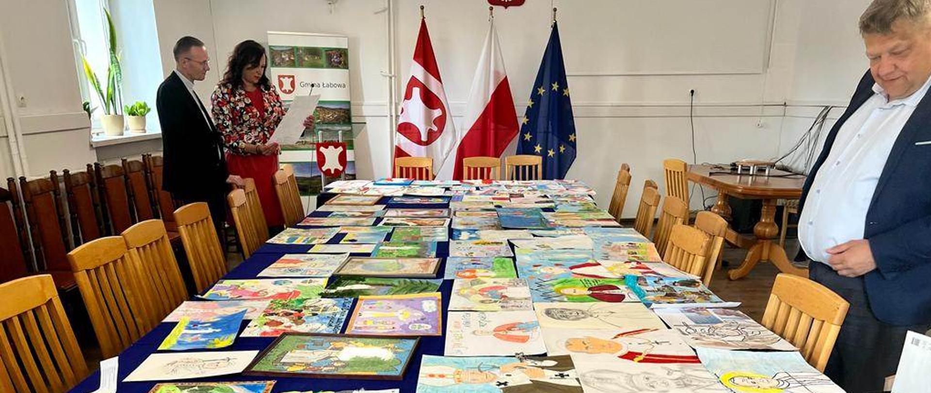 Na stole leżą prace konkursowe o Janie Pawle II, komisja konkursowa ogląda prace