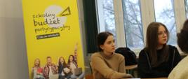 Uczennice siedzą obok baneru promującego pilotażowy program wdrażania szkolnego budżetu