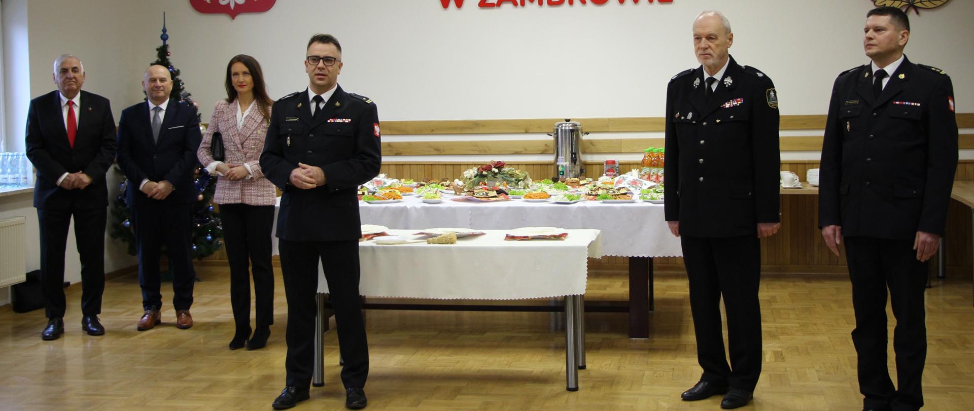 Zdjęcie przedstawia świąteczne spotkanie opłatkowe w sali z napisem "KOMENDA POWIATOWA PAŃSTWOWEJ STRAŻY POŻARNEJ W ZAMBROWIE" oraz godłem Polski na ścianie. Na tle stołu przykrytego białym obrusem stoi grupa osób: od lewej Burmistrz Miasta Zambrów, Wójt Gminy Zambrów, Starosta Powiatu Zambrowskiego, Zastępca Podlaskiego Komendanta Wojewódzkiego PSP, Senator RP Marek Adam Komorowski oraz Komendant Powiatowej PSP w Zambrowie.