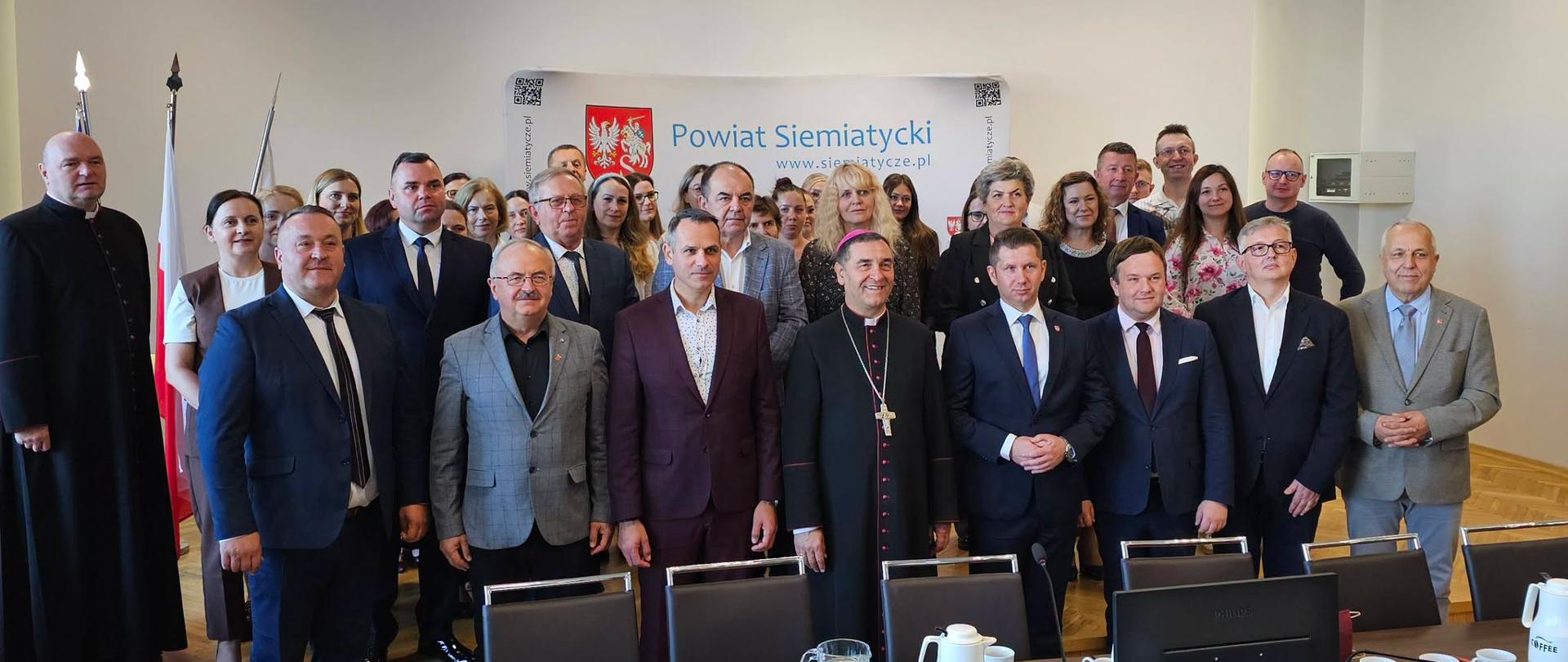 Duża grupa osób, w tym mężczyźni i kobiety w strojach galowych, stoi razem do zdjęcia w sali konferencyjnej, a w tle widać flagi i transparenty.