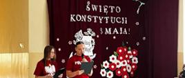 Dwie uczennice ubrane w bordowe koszulki z wizerunkiem białego orła i długi czarne spódnice przemawiają do mikrofonu, w tle dekoracja z biało-czerwonych kwiatów oraz napis "Święto konstytucji 3 maja"