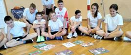Uczniowie w strojach sportowych biały podkoszulek ciemne krótkie spodenki siedzą na podłodze obok bramki piłkarskiej i prezentują położone na podłodze książki o sportowcach