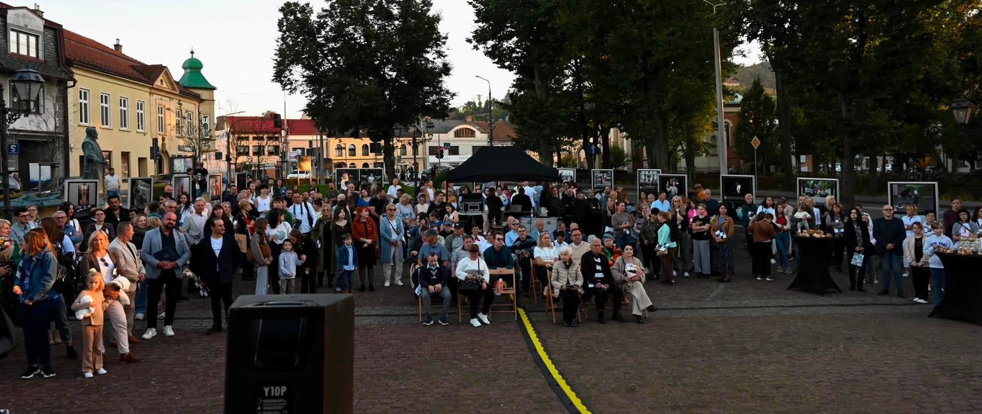 Zdjęcie przedstawia uczestników wydarzenia zgromadzonych na rynku w Kalwarii Zebrzydowskiej. W centrum widoczna jest liczna grupa mieszkańców stojących i siedzących przed sceną, a wokół placu ustawiono plansze ze zdjęciami tworzące plenerową ekspozycję. W tle znajdują się zabytkowe kamienice, drzewa oraz zabudowa centrum miasta.