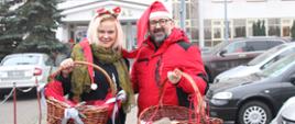 Zdjęcie przedstawia Kierownik Referatu Promocji Powiatu Przasnyskiego wraz ze Starostą Przasnyskim na parkingu przasnyskiego szpitala. W dłoniach trzymają kosze z prezentami dla dzieci, w tle budynek przasnyskiego szpitala.