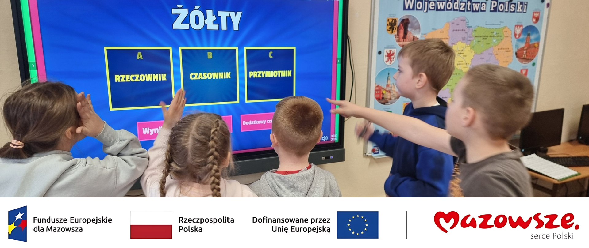 Grupa pięciorga dzieci stoi przodem do dużego ekranu interaktywnego, rozwiązując quiz językowy. Na niebieskim ekranie widoczne jest słowo "ŻÓŁTY" oraz trzy opcje do wyboru: A - RZECZOWNIK, B - CZASOWNIK, C - PRZYMIOTNIK. Dwoje dzieci po prawej stronie wskazuje rękami na ekran. W tle wisi mapa przedstawiająca "Województwa Polski". Na dole zdjęcia znajduje się biały pasek z logotypami Funduszy Europejskich dla Mazowsza, Rzeczypospolitej Polskiej, Unii Europejskiej oraz marki Mazowsze serce Polski.