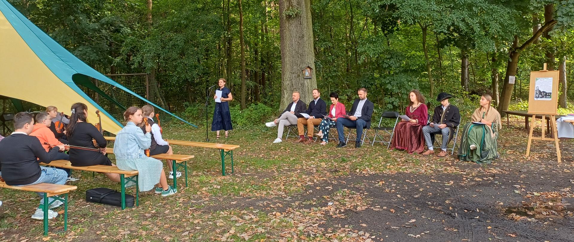 Ludzie siedzący na ławkach pod dużym drzewem. W tle las.