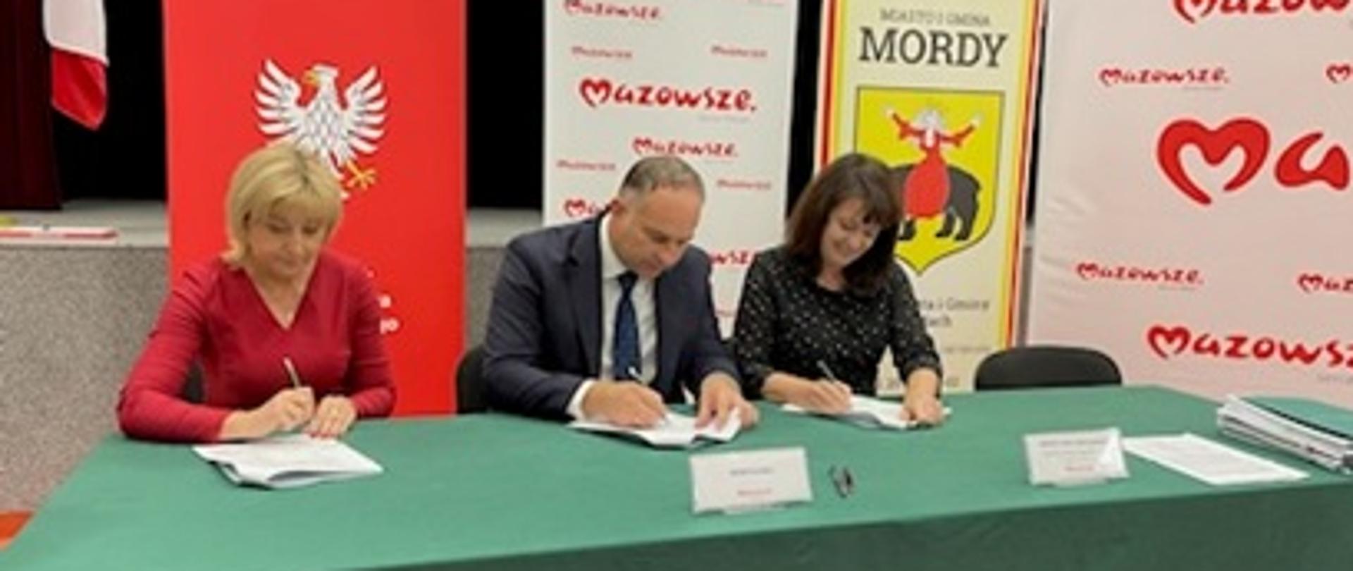 przy stole przykrytym zielonym materiałem siedzą trzy osoby, w czasie podpisywania umowy
