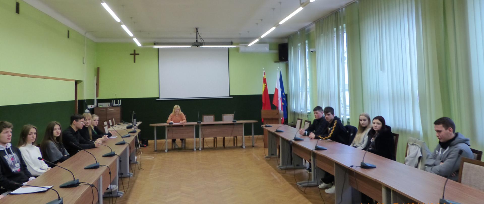 Członkowie Młodzieżowej Rady Powiatu Proszowickiego podczas sesji obradują przy stolach, na których znajdują się mikrofony. W tle zielona lamperia oraz stojak z flagami Polski, Unii Europejskiej i Powiatu Proszowickiego.