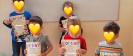 Na zdjęciu pięcioro dzieci z zasłoniętymi twarzami stoi przed tablicą i trzyma dyplomy. W górnej części zdjęcia znajduje się napis „Wspaniały Świat Literatury” oraz ilustracje książek. W dolnej części, przed dziećmi, ustawiono kolorowe torby z upominkami. 