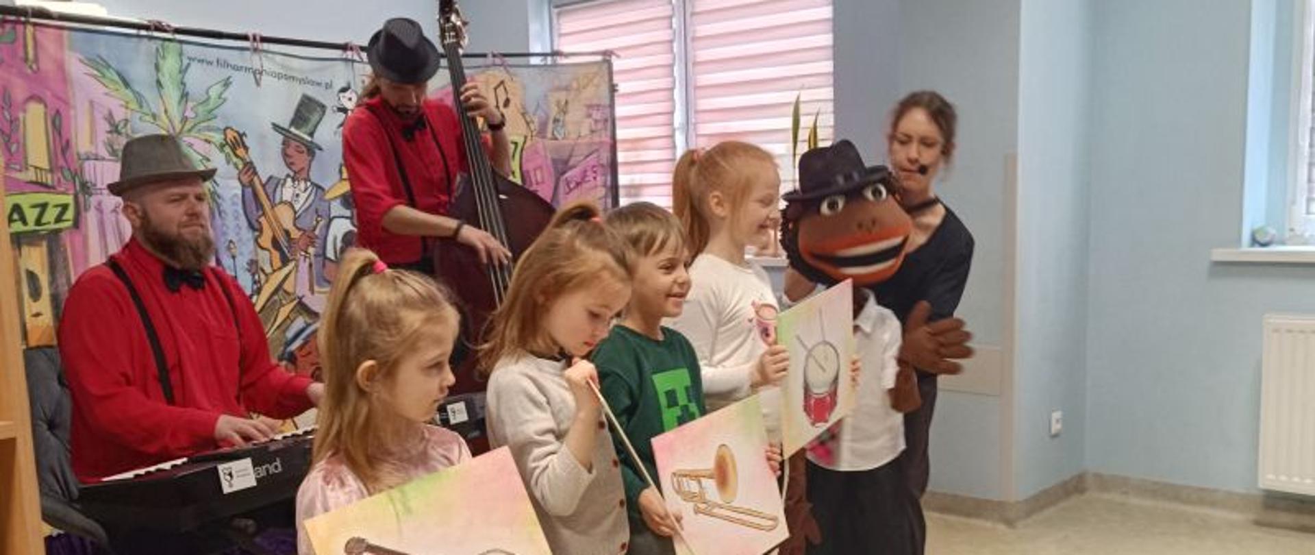 Czworo dzieci w wieku przedszkolnym stoi w rzędzie trzymając w rękach rysunki przedstawiające instrumenty muzyczne. Za nimi dwóch mężczyzn w czerwonych koszulach i czarnych kapeluszach na głowie. Jeden z nich siedzi przed organami, drugi trzyma kontrabas. Obok dzieci stoi kobieta trzymająca przed sobą dużą lalkę. Za nimi tło przedstawiające muzyków a po prawej stronie okno.