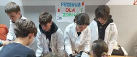 Grupa uczniów w białych fartuchach medycznych pomaga młodszym kolegom w wykonaniu pomiaru ciśnienia tętniczego przy stanowisku z plakatem „PRZERWA DLA ZDROWIA”. W tle widać zaangażowanych asystentów pochylonych nad aparaturą, podczas gdy na pierwszym planie dziewczynka z jabłkiem w dłoni czeka na wynik badania.