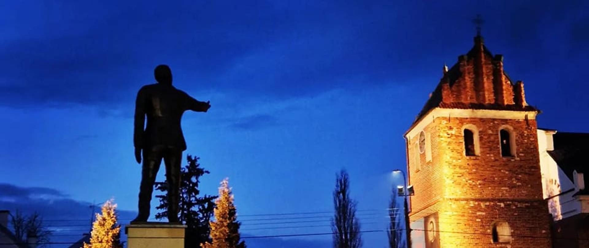 Zdjęcie wykonane o świcie przedstawia rzeźbę burmistrza Matuszewskiego oraz dzwonnicę kościoła farnego w Przasnyszu.