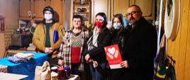 Zdjęcie przedstawia Starostę Krzysztofa Bieńkowskiego wraz z Panią Haliną oraz wolontariuszami Szlachetnej Paczki. Fotografia wykonana w domku Pani Haliny, na stole obok przywiezionych darów znajdują się wykonane przez Panią Halinę w odwdzięce przetwory oraz wełniane czapki i rękawiczki. 