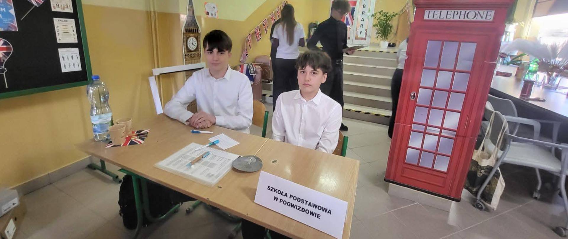 Dwóch chłopców w białych koszulach siedzi przy ławce szkolnej. W tle widać różne charakterystyczne dla Wielkiej Brytanii repliki budowli i symboli.