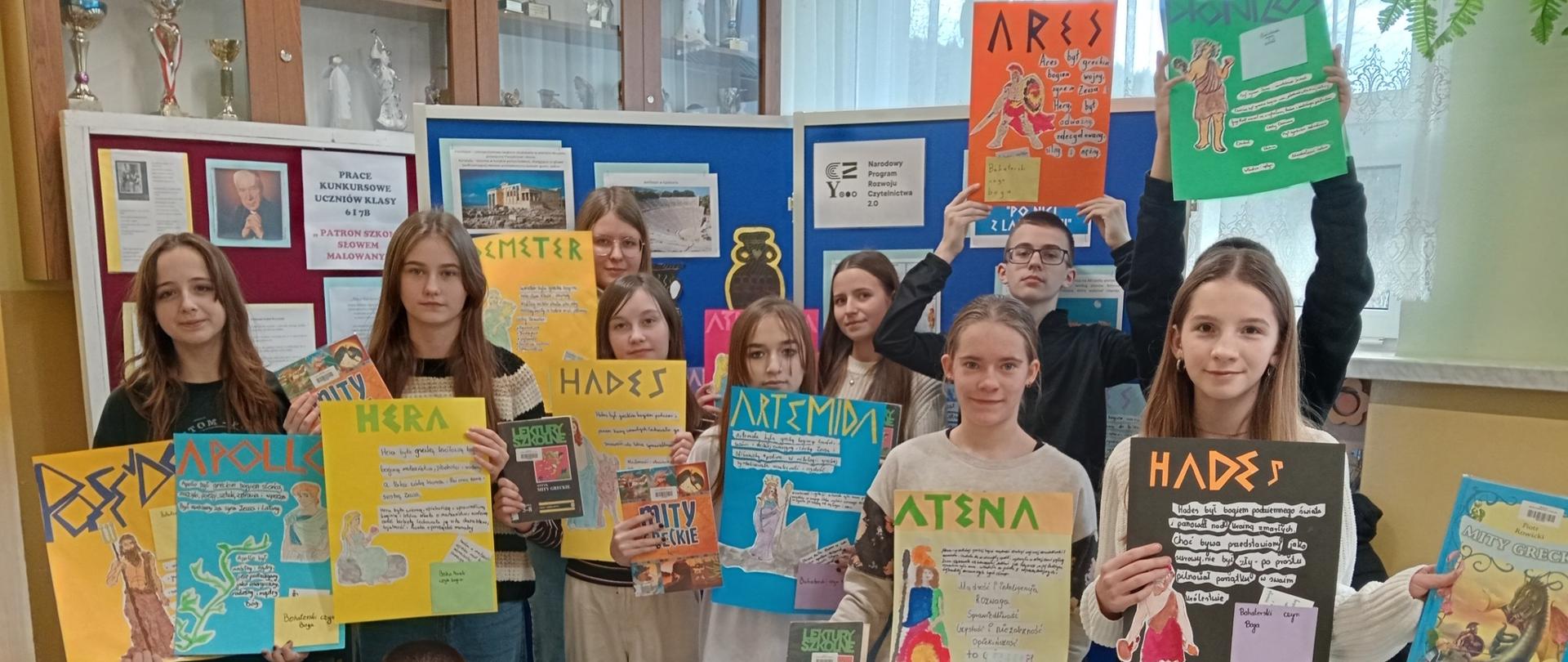 Uczniowie klasy 7 trzymają w ręku wykonane przez siebie kolorowe plakaty z greckimi bogami i herosami oraz książki traktujące o tematyce greckich bogów