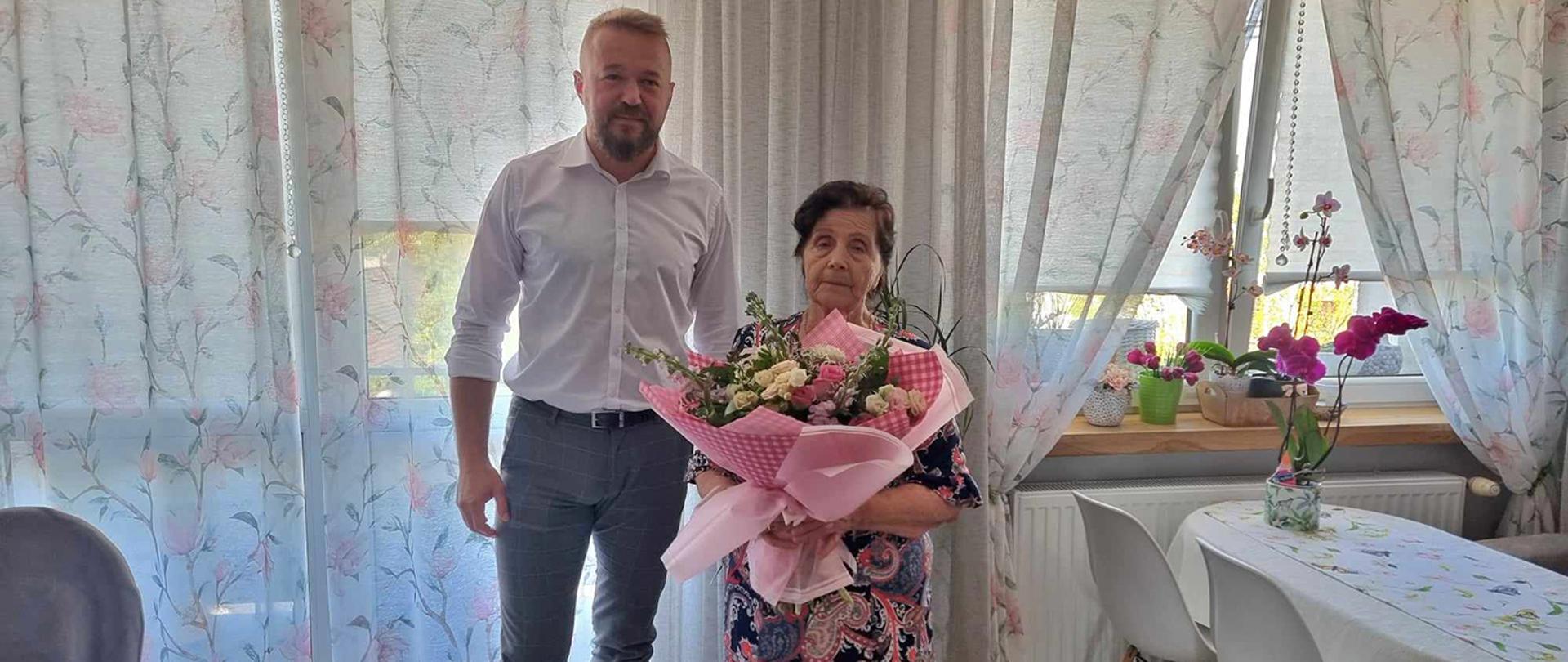 Mężczyzna w białej koszuli obok starsza Pani z bukietem kwiatów. W tle jasne zasłony.