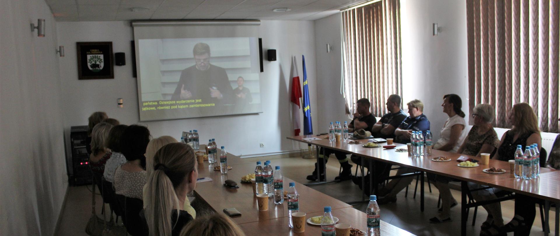 Uczestnicy konferencji siedzą przy biurku, przy monitorze zdalnym