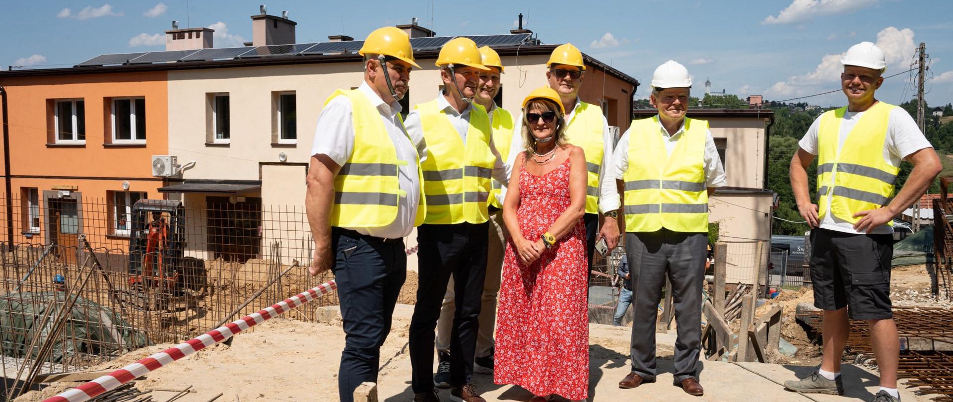 Na zdjęciu widać grupę siedmiu osób stojących na placu budowy w słoneczny dzień. Sześciu mężczyzn ubranych jest w żółte kamizelki odblaskowe, a czterech z nich ma na głowie żółte kaski ochronne, dwóch – białe kaski. W środku grupy stoi kobieta w długiej, czerwonej sukience w białe wzory, bez kasku i kamizelki. W tle widoczne są dwukondygnacyjne budynki w odcieniach pomarańczowym i kremowym, fragmenty konstrukcji budowlanej oraz sprzęt budowlany. Pogoda jest słoneczna, niebo niemal bezchmurne.