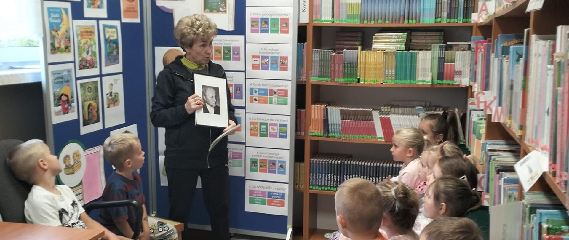 Uczniowie klasy 1 siedzą na ławeczkach w bibliotece, słuchają pani bibliotekarki, która opowiada o autorce :Szewczyka Dratewki" pisarce Janinie Porazińskiej, za bibliotekarką stoi ścianka moderacyjna, na której zamieszczone zostały okładki książek napisanych przez Porazińską oraz quiz ze znajomości lektury "Szewczuk Dratewka"