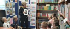 Uczniowie klasy 1 siedzą na ławeczkach w bibliotece, słuchają pani bibliotekarki, która opowiada o autorce :Szewczyka Dratewki" pisarce Janinie Porazińskiej, za bibliotekarką stoi ścianka moderacyjna, na której zamieszczone zostały okładki książek napisanych przez Porazińską oraz quiz ze znajomości lektury "Szewczuk Dratewka"