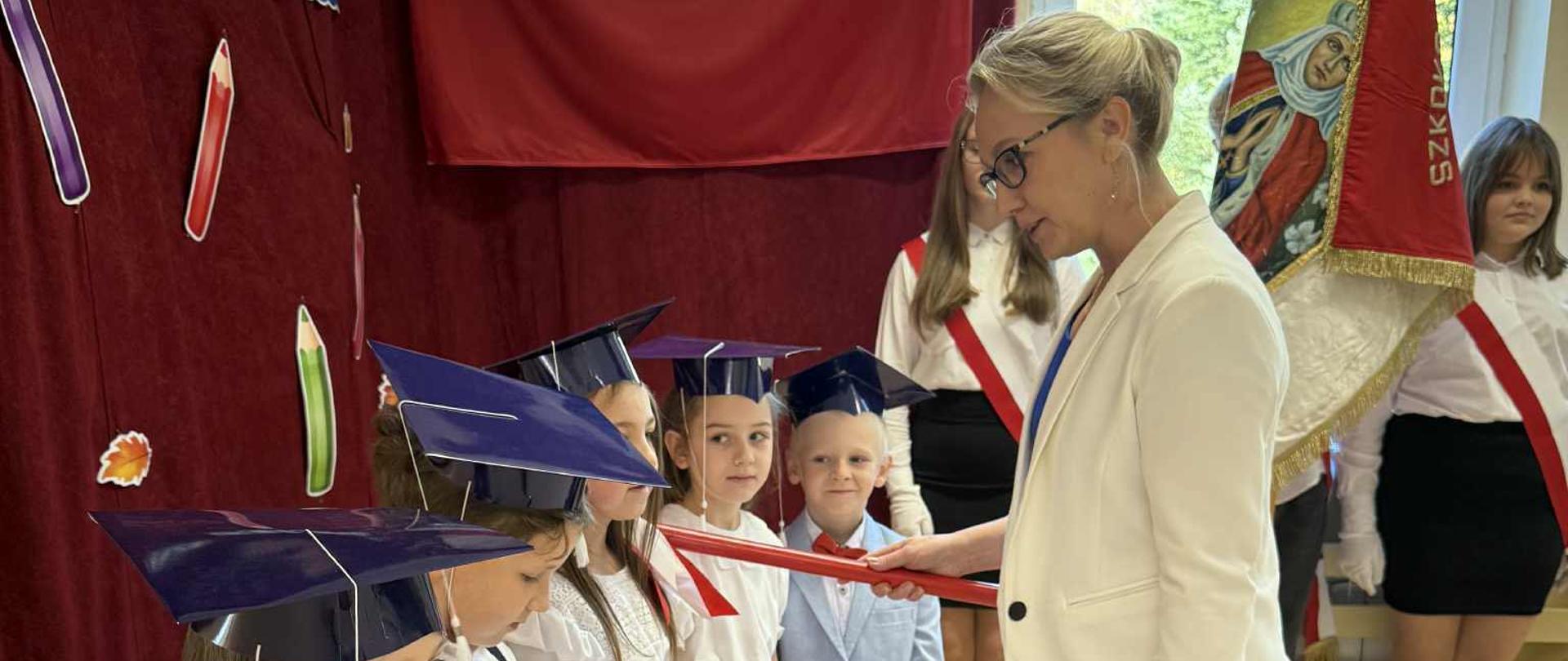 Pani dyrektor pasuje czerwonym ołówkiem odświętnie ubranych uczniów klasy 1