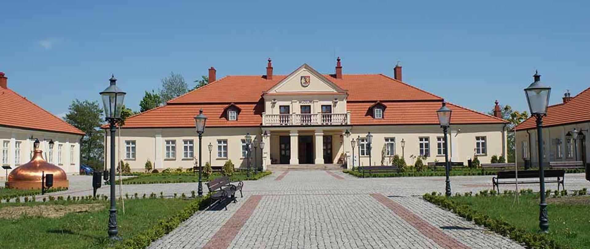 Duży, dwupiętrowy beżowy budynek, pokryty czerwona dachówką. Przed budynkiem ułożona kostka, ustawione latarnie i ławki. 