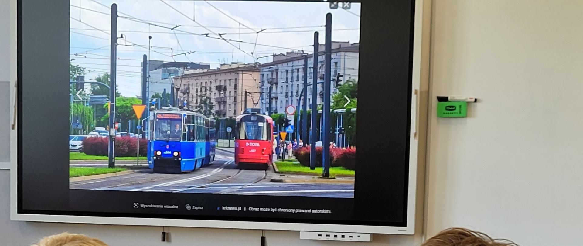 Na górze zdjęcia ekran w jasnej ramce przedstawiający dwa mijające się tramwaje, jeden w kolorze czerwonym, drugi w kolorze niebieskim. Poniżej dwie sylwetki osób siedzące tyłem, jedna na zielonym krześle, druga na żółtym. Pomiędzy nimi fragment szarego stołu, z bordową bluzą leżącą na górze.