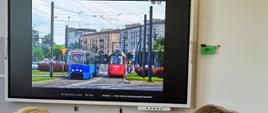 Na górze zdjęcia ekran w jasnej ramce przedstawiający dwa mijające się tramwaje, jeden w kolorze czerwonym, drugi w kolorze niebieskim. Poniżej dwie sylwetki osób siedzące tyłem, jedna na zielonym krześle, druga na żółtym. Pomiędzy nimi fragment szarego stołu, z bordową bluzą leżącą na górze.