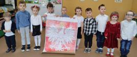 Chłopcy i dziewczynki z oddziału przedszkolnego trzymają duże narysowane na kartonie godło Polski przedstawiające orła w złotej koronie na czerwonym tle