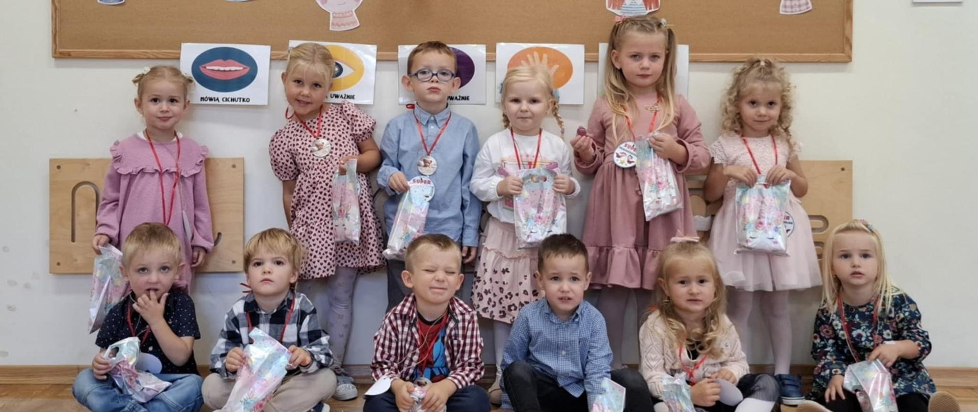 Grupa dzieci radosnych i uśmiechniętych z papierowymi medalami siedzi po turecku. W tle tablica korkowa z kolorowym napisem "Dzień Przedszkolaka".