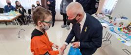 Starosta Kraśnicki Andrzej Rolla składa gratulacje laureatowi konkursu "Bezpieczna Zima 2022", ściska dłoń młodego chłopca i wręcza niebieską torebkę nagród. 