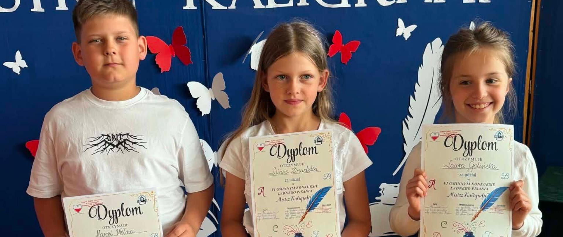 Na zdjęciu widoczna jest trójka uczniów stojących na tle granatowej dekoracji przygotowanej z okazji konkursu kaligraficznego. Każde z dzieci trzyma w rękach dyplom potwierdzający udział i osiągnięcia w konkursie. W tle można odczytać napis informujący o konkursie ładnego pisania „Mistrz Kaligrafii”, a dekorację uzupełniają papierowe pióra i motyle.
Uczniowie pozują do pamiątkowego zdjęcia po uroczystym wręczeniu nagród. Dziewczynki ubrane są odświętnie w spódniczki i jasne bluzki, natomiast chłopiec ma na sobie białą koszulkę i jasne spodenki. Wszyscy prezentują otrzymane dyplomy, będące efektem ich starannej pracy i umiejętności pięknego pisania.