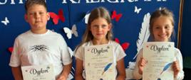 Na zdjęciu widoczna jest trójka uczniów stojących na tle granatowej dekoracji przygotowanej z okazji konkursu kaligraficznego. Każde z dzieci trzyma w rękach dyplom potwierdzający udział i osiągnięcia w konkursie. W tle można odczytać napis informujący o konkursie ładnego pisania „Mistrz Kaligrafii”, a dekorację uzupełniają papierowe pióra i motyle.
Uczniowie pozują do pamiątkowego zdjęcia po uroczystym wręczeniu nagród. Dziewczynki ubrane są odświętnie w spódniczki i jasne bluzki, natomiast chłopiec ma na sobie białą koszulkę i jasne spodenki. Wszyscy prezentują otrzymane dyplomy, będące efektem ich starannej pracy i umiejętności pięknego pisania.