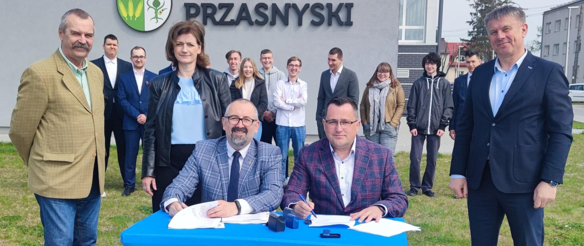 Zdjęcie przedstawia moment podpisania umowy na budowę basenów odkrytych w powiecie przasnyskim przy Zespole Szkół Powiatowych przy ulicy Mazowieckiej w Przasnyszu. Przy niebieskim stoliku siedzą Starosta Przasnyski Krzysztof Bieńkowski oraz przedstawiciel wykonawcy, firmy Rembud. Po prawej przedstawiciel firmy Rembud, po lewej Wicestarosta Waldemar Trochimiuk oraz Skarbnik Beata Iwona Czaplicka, w tle młodzież szkolna.