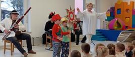 Zdjęcie prezentuje dzieci biorące udział w audycji muzycznej oraz teatrzyku. Przed grupą siedzących dzieci występują aktorzy