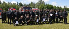 Duże zdjęcie grupowe wszystkich uczestników zawodów pożarniczych na zielonej murawie. Kilkudziesięciu strażaków w mundurach galowych i bojowych pozuje razem z cywilami na tle zaparkowanych czerwonych wozów strażackich. Niektórzy z nich trzymają puchary i dyplomy.