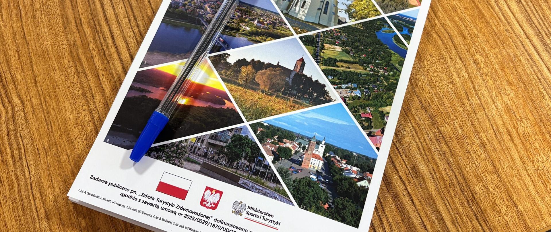 Notes/broszura projektu "Szkoła Turystyki Zrównoważonej" leżąca na drewnianym stole, z niebieskim długopisem na okładce. Okładka to kolaż zdjęć krajobrazów i architektury, zawierający logo Ministerstwa Sportu i Turystyki oraz informację o dofinansowaniu.