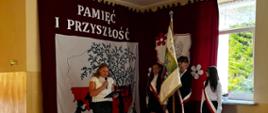 Uczennica z klasy 6 ubrana na galowo przemawia do mikrofonu podczas akademii, w tle dekoracja z napisem "Pamięć i przyszłość", po prawej poczet sztandarowy szkoły