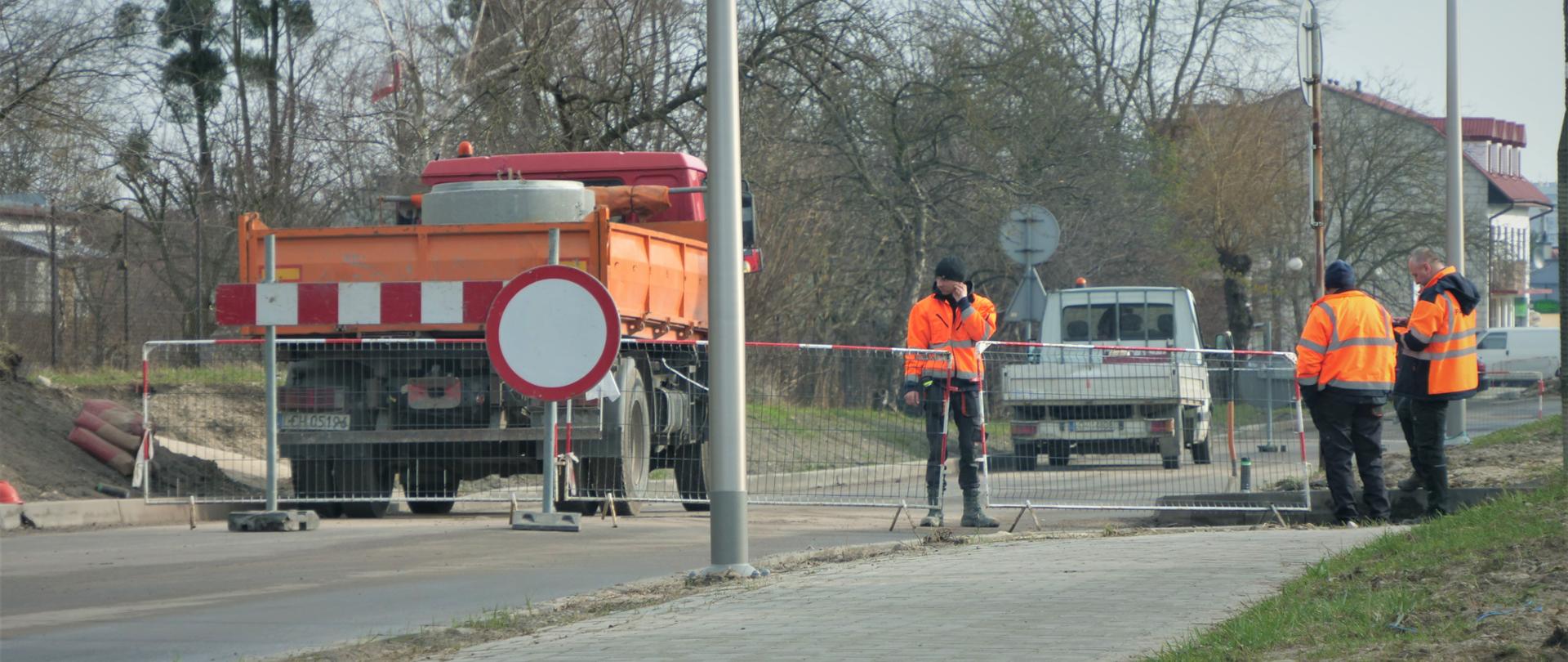 Zdjęcie przedstawia prace drogowe. Na drodze stoi samochód ciężarowy, a przed nim barierki oraz znak zakaz wjazdu. Za barierkami stoją ludzie ubrani w pomarańczowe kamizelki odblaskowe.